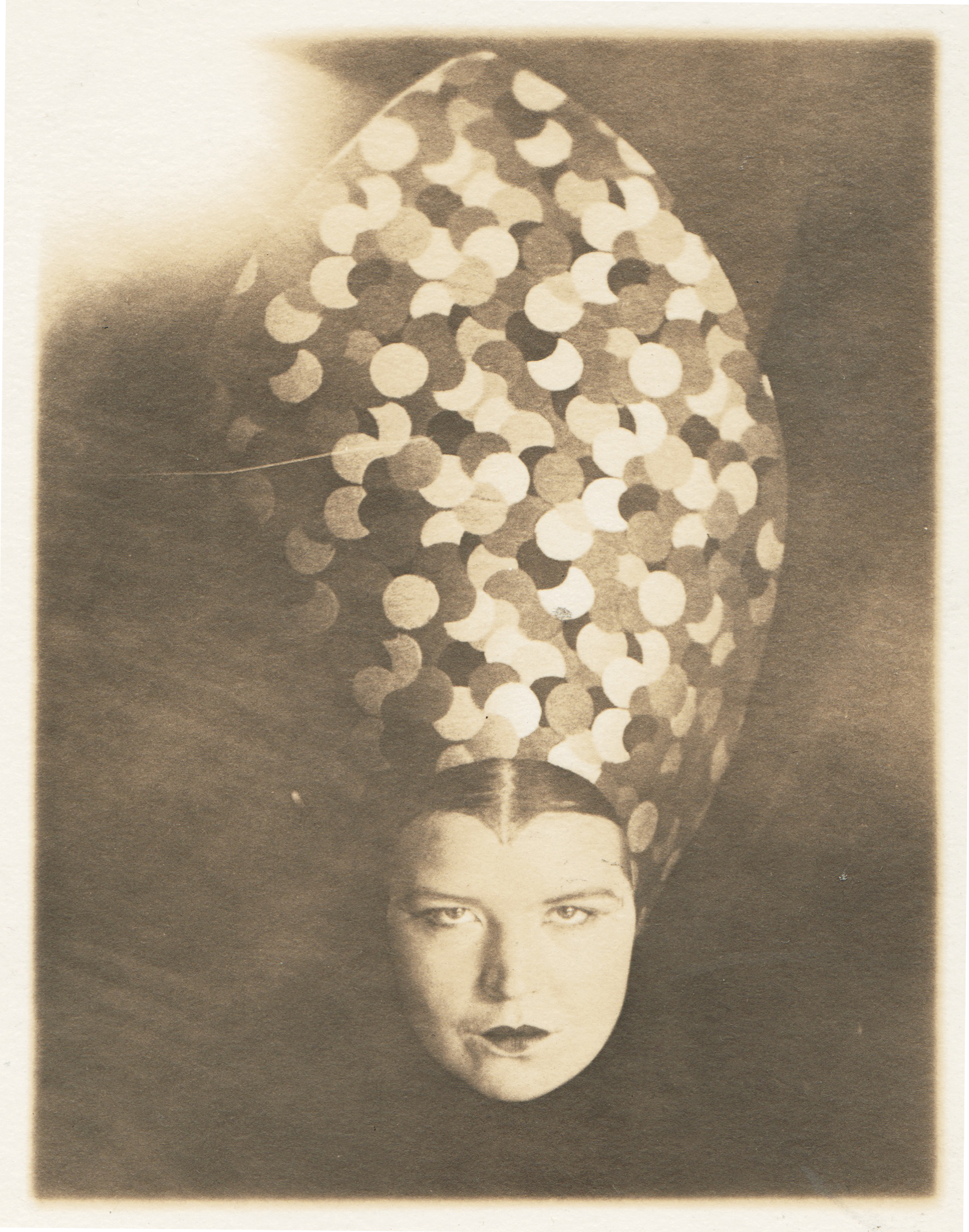 a black and white photograph, possibly from the 1920s or 1930s, depicts a woman wearing a striking, avantgarde hat. the hat is a pointed, ovular shape completely covering her head and extending down to her shoulders. it's decorated with numerous small, overlapping circles in varying shades of gray and beige, creating a geometric pattern. the woman's face is partially visible, with her expression appearing serious and somewhat mysterious. the background is a plain dark tone which puts emphasis on the woman and her hat. the overall style suggests an art deco influence. the photo has a slightly aged, vintage quality.