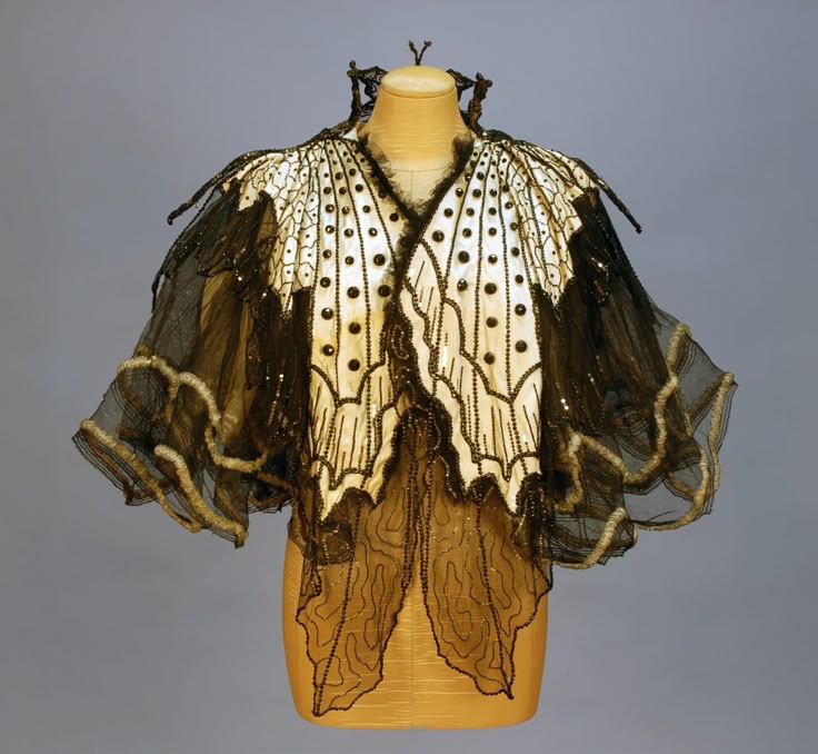 a butterflyshaped cape is displayed on a mannequin. the cape is mostly white with black beaded accents and a black netlike fabric hanging off the edges. the silhouette of the garment is reminiscent of butterfly wings.