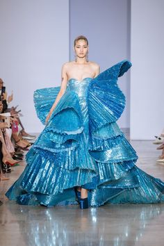 a female model with long black hair and wearing a bright blue gown with pleating throughout poses on the runway. the dress has several tiers and is embellished with glitter. the model has a natural makeup look, with a light lip, a small amount of eyeliner, and nude eyeshadow. she is looking forward and appears confident. there are other people in the background, though they are out of focus.