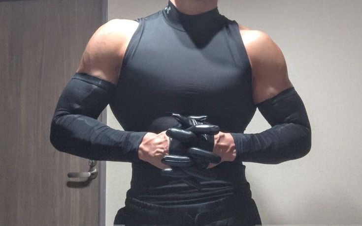 he image shows a muscular man with a welldefined physique wearing a black sleeveless shirt and black arm sleeves. he is holding black gloves in his hands. the background is a simple grey wall and a part of a brown door is visible. the man's arms and shoulders are prominently displayed, highlighting his muscular build. the overall impression is of strength, fitness, and athleticism.