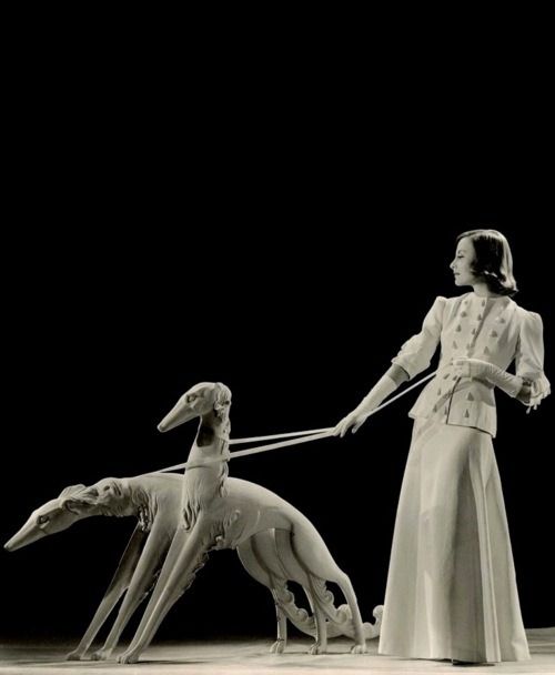 a black and white vintage portrait of a woman in a long white dress walking two greyhounds on leashes. the woman is posed in a classic fashion pose with her hands on her hips, and the greyhounds are walking beside her. the background is black, and the photo is taken in a studio setting.