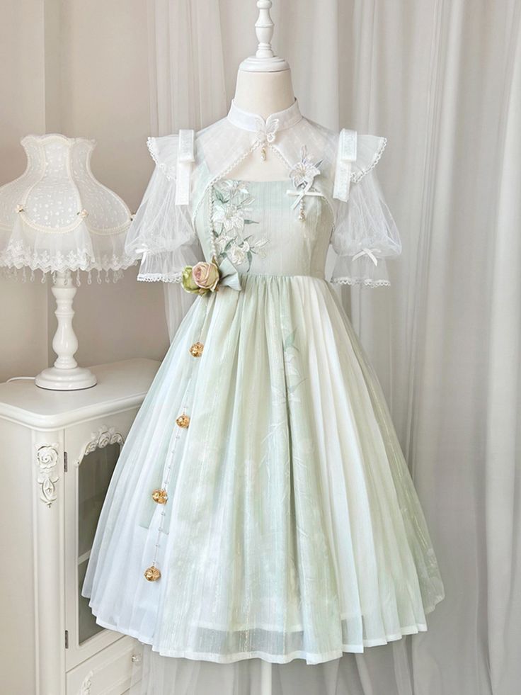 a fulllength photo of a light green and white lolita fashion dress. the dress has a high neckline, sheer sleeves, and floral accents. it is displayed on a mannequin in front of a white curtain.  there's a white nightstand and a white lamp to the left of the dress.