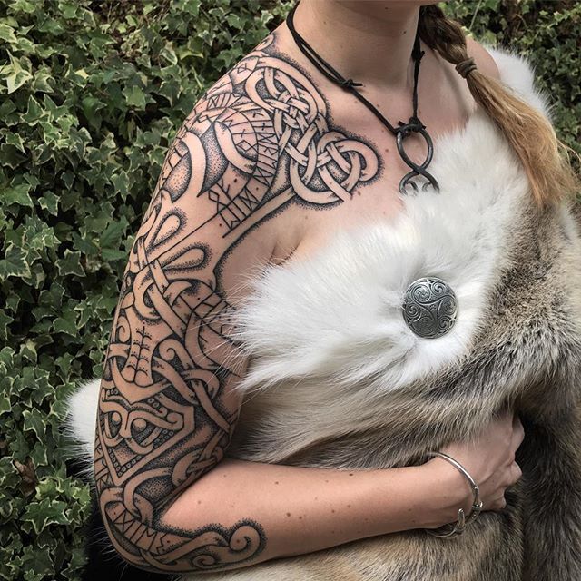 a woman is shown from the chest up, facing forward, wearing a fur coat and jewelry. her right arm and shoulder are heavily tattooed in a vikinginspired style, featuring celtic knots and runes. she is wearing a black cord necklace with a pendant and a silver bracelet on her right wrist. her blonde hair is styled in braids. a large, round silver brooch secures the fur coat.