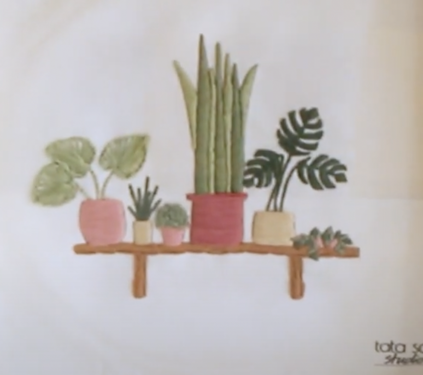 the image showcases a piece of modern embroidery on a white fabric. it depicts a wooden shelf with five potted plants, including a snake plant, a monstera leaf, and a succulent. the plants are stylized and stitched with various shades of green thread, while the pots are pink and beige.