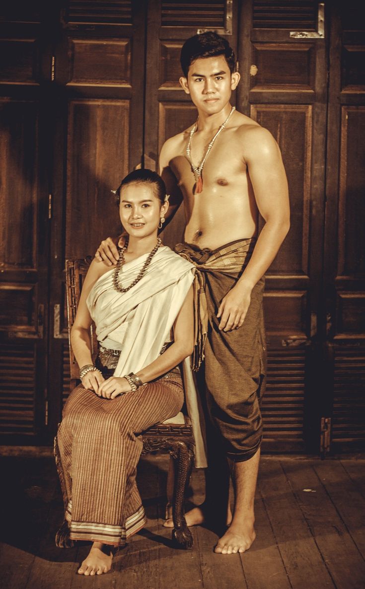 a sepiatoned portrait of a young thai couple dressed in traditional clothing. the woman is seated on an antique wooden chair wearing a light beige shawl and a brown striped sarong. she wears several bracelets and a pearl necklace. the man stands behind her, shirtless except for a brown sarong wrapped around his waist. he also wears a necklace. they are posed against a background of wooden doors, giving a vintage or historical feel to the image. the overall mood is elegant and romantic.
