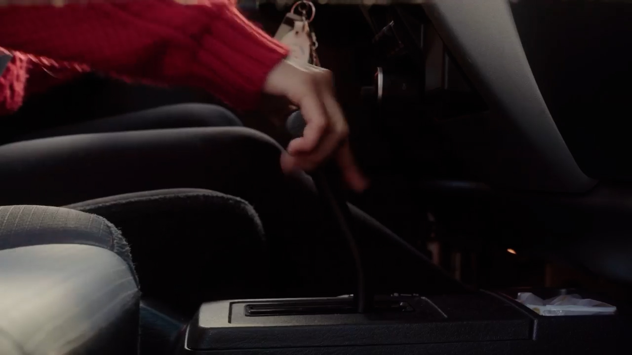 a closeup shot shows a woman's hand shifting gears in a car with a manual transmission. the focus is on the gear shift and the woman's hand, with the rest of the car interior blurred in the background. the lighting suggests a bright, sunny day, with the sunlight filtering through the car windows. the overall color palette is muted and warm, with shades of red, brown, and black.