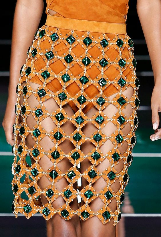 loseup of a woman's model wearing a short, orange, cutout skirt embellished with numerous darkgreen gemstones. the skirt is intricately designed with a gridlike pattern created by the cutouts and the gemstones. the model's skin tone is visible through the openings in the skirt. the background is blurred, suggesting a fashion show or runway setting. the overall impression is one of high fashion and luxury.
