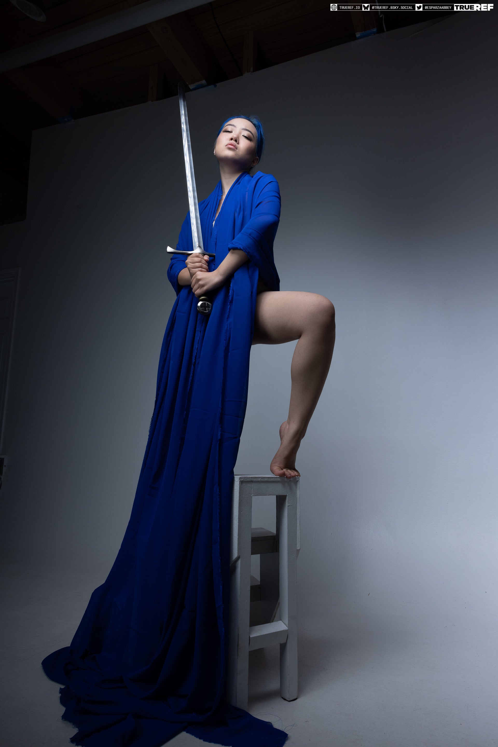 a woman with blue hair and a long blue robe is standing with one foot on a white stool, holding a sword and looking upwards in a studio setting.