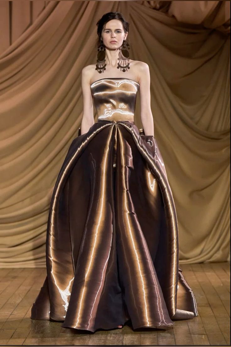 a model in a flowing brown gown with gold accents walks down a runway with a backdrop of tan curtains. the gown is strapless with a fitted bodice and a full skirt. the model is wearing large statement earrings and her hair is pulled back. she looks very elegant and glamorous. the dress has a sophisticated and classic silhouette. the image is taken from a high fashion runway show.