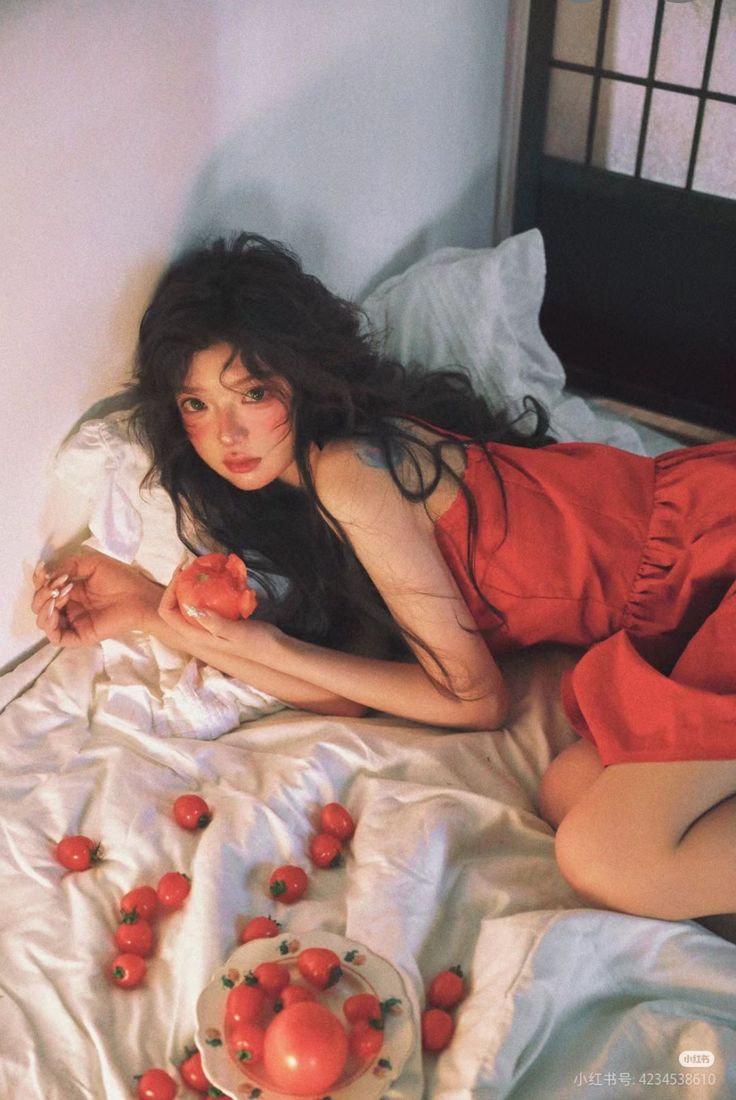 a woman with long, dark hair is lying on a bed covered in a white sheet. she's wearing a red dress and holding a tomato. a plate of tomatoes and scattered single tomatoes are also on the bed. the woman has fair skin with pink blush on her cheeks and her hair is styled in loose waves. she has a tattoo on her left shoulder. a white pillow is behind her head. a window with a grid pattern is visible in the background.