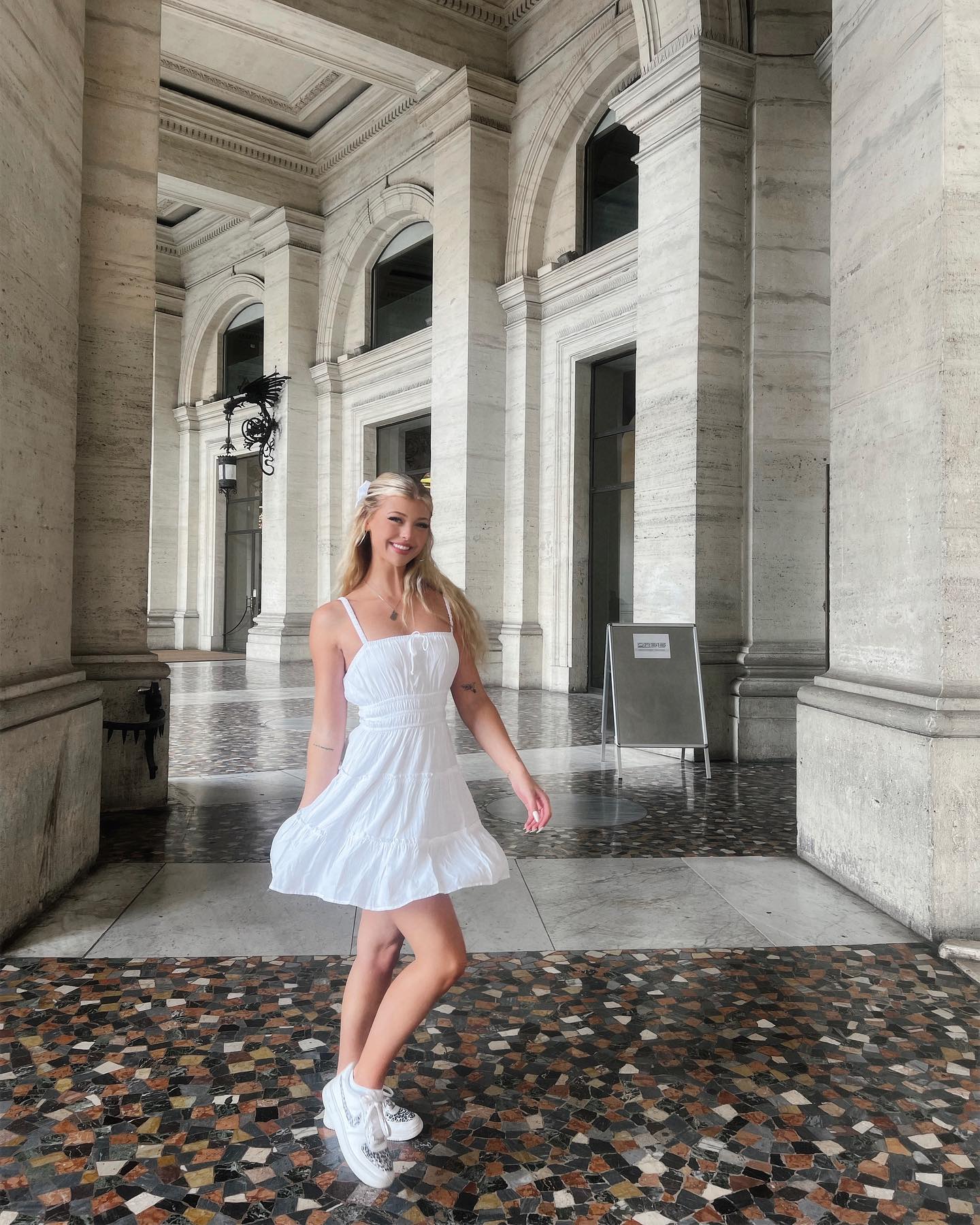 a young blonde woman in a white dress and sneakers stands in front of a building with tall columns. she is smiling and holding the edge of her dress as if walking. the building has arched doorways and windows, and the floor is tiled. the light is bright and sunny.