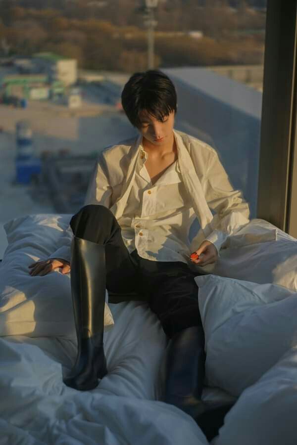 a young man with dark hair, wearing a loose, partially unbuttoned white shirt, black pants, and black riding boots, is sitting on a bed with white linens. he is positioned in front of a large window, looking down with a thoughtful expression. the window offers a view of a cityscape bathed in warm, afternoon sunlight. the overall atmosphere is relaxed and intimate.
