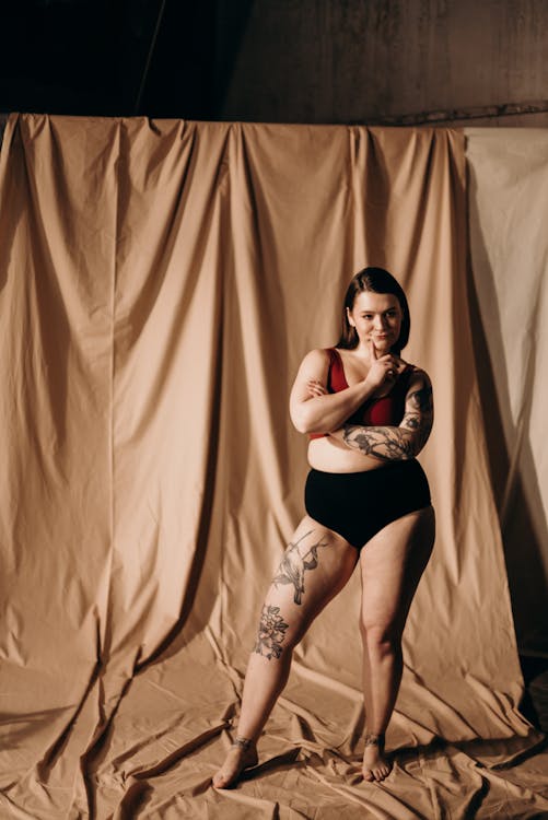 a plussize woman with tattoos stands confidently against a beige backdrop, her arms crossed, wearing black underwear and a red bra. she projects selfassuredness and comfort in her own skin, embodying body positivity. the studio setting enhances the focus on the model and her expression.