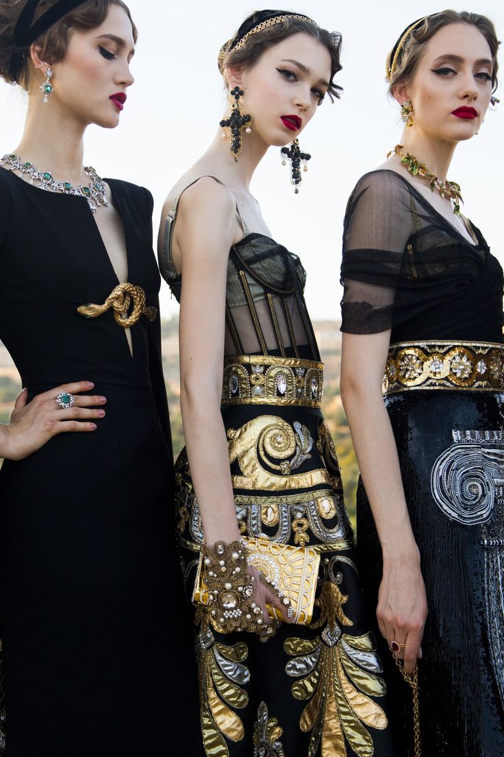 hree models are posing outdoors in elaborate dolce & gabbana alta moda gowns.  the dresses are predominantly black with intricate gold embroidery and embellishments.  the models wear ornate jewelry, including large earrings and necklaces, and carry clutches. their hair is styled in elegant updos, and they are wearing bold red lipstick.  the setting appears to be a sunny outdoor location, possibly with a mediterranean vibe. the overall aesthetic is one of high fashion, luxury, and glamour.