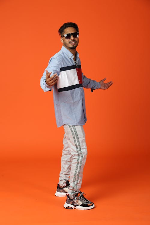 a man standing in front of an orange background, wearing sunglasses, a blue shirt with a white and red square on the chest, light gray pants with a green stripe down the side, and multicolored sneakers.
