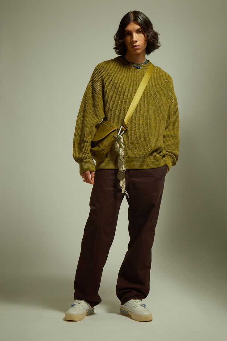 a fullbody studio shot of a male fashion model. he is wearing a loosefitting, olive green ribbed sweater, dark brown straightleg pants, and offwhite chunky sneakers. he carries a small olive green crossbody bag with a long strap. his long brown hair is styled naturally. the background is a plain, light beige. the overall style is casual and relaxed, indicative of streetwear or autumn/fall fashion.