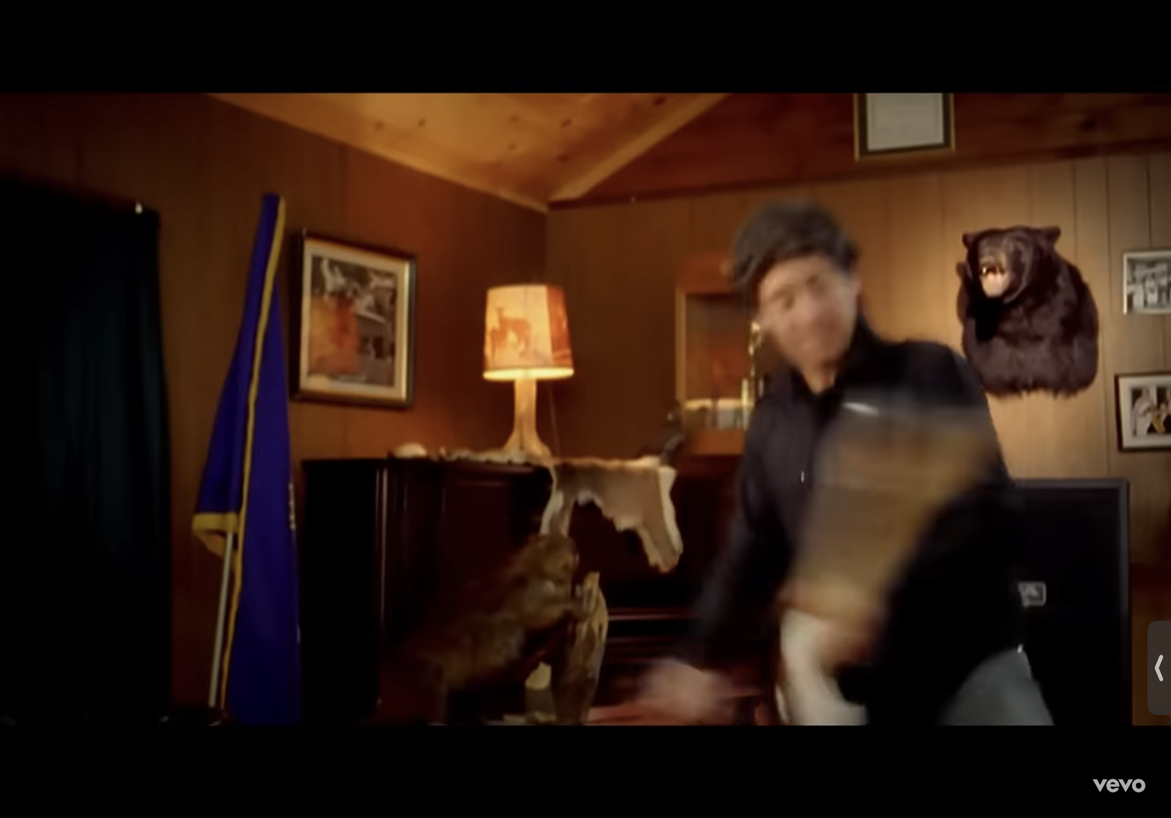 the image captures a man in midmotion, seemingly dancing enthusiastically while holding a guitar. he is in a rustic, woodpaneled room adorned with a mounted bear head, a flag, and westernthemed pictures. the warm, artificial lighting casts a cozy glow on the scene, emphasizing the man's energetic movement.