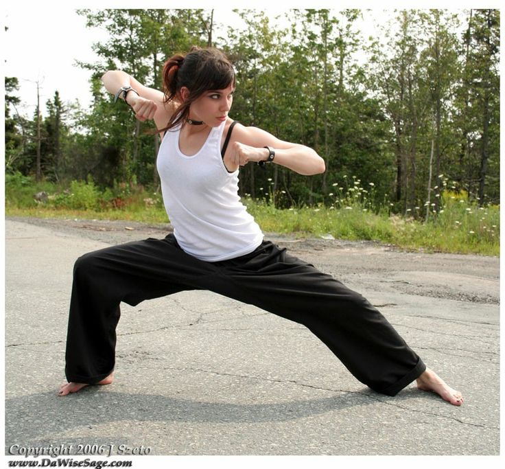 a young woman dressed in black pants and a white tank top is doing a martial arts pose on a country road. she is looking off to the side with a serious expression on her face and she looks like she's about to strike.  the road is surrounded by grassy areas and trees, and it looks like a sunny summer day.