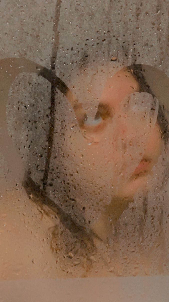 the image shows a closeup of a woman's face, partially obscured by steam and water droplets on a shower glass.  the details of her face are blurred and softened by the condensation, creating a hazy, intimate, and mysterious mood. the overall tone is warm and calm, suggestive of a relaxing shower or spa experience.  the focus is on the ethereal quality of the image, rather than sharp details.