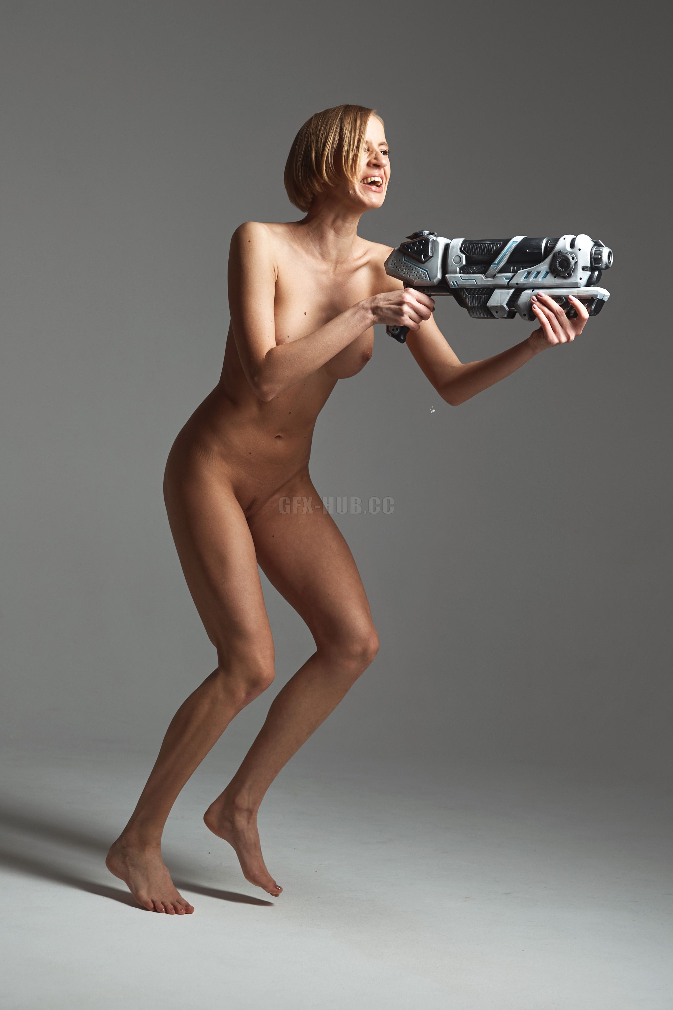 a nude woman with fair skin and short blonde hair is running while holding a futuristic gun in her hands. she is laughing and appears to be in motion. the background is a solid gray color.