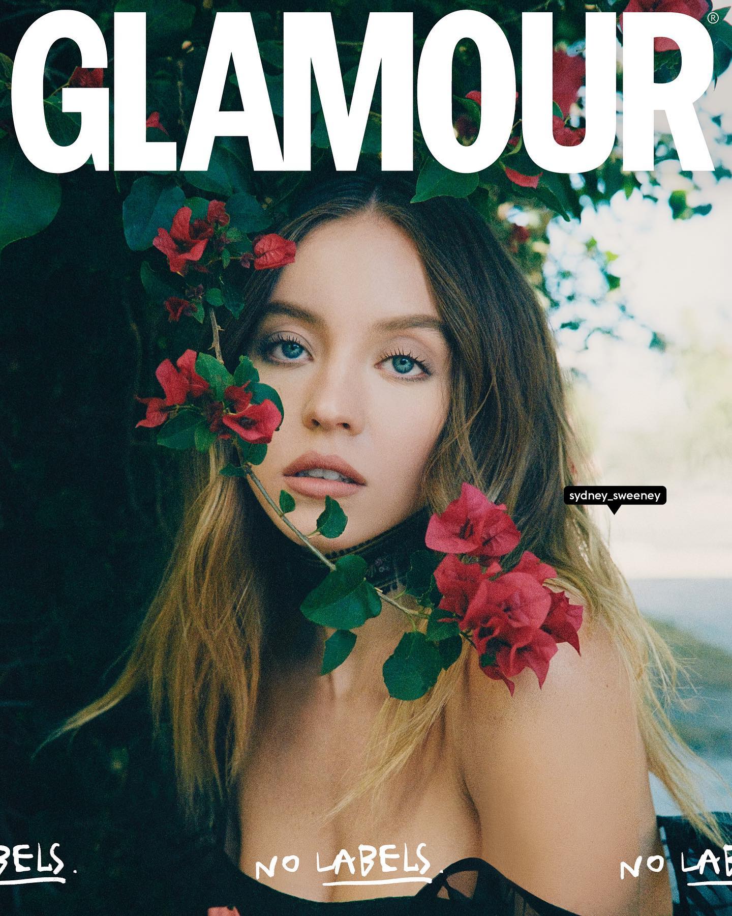the image is a magazine cover for glamour, featuring actress sydney sweeney.  sweeney is the central focus, her face and upper body visible. she's posed with bougainvillea flowers draped around her shoulders and neck. the background is blurred, showing more greenery. the text "glamour" is prominently displayed at the top of the image, and "no labels" is written in a handwritten style across the bottom.  the overall style is chic and natural, emphasizing sweeney's beauty. the image has a warm tone and a slightly vintage feel.