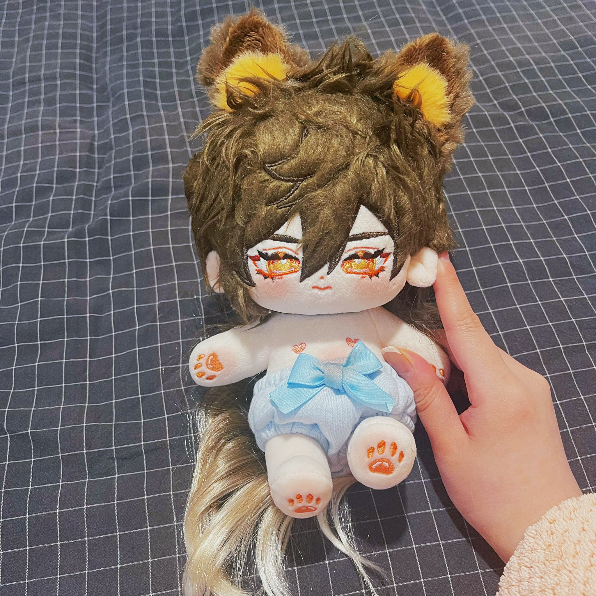 a plush doll with brown hair and yellow ears wearing a blue diaper, being held by a hand.  the doll is laying on a black and white checkered fabric.  the doll is very cute and soft.
