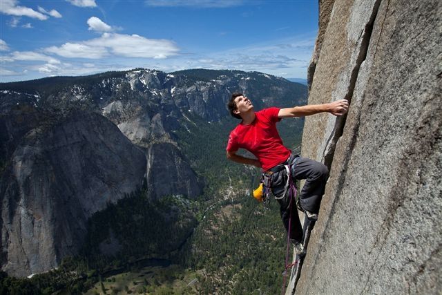 a rock climber scales a sheer rock face, looking up at the summit. the climber is secured with a rope and harness, but is climbing free solo, without any additional aid. the background shows the vast landscape of yosemite national park with el capitan and other peaks in the distance. the climber is dwarfed by the sheer scale of the rock face,  highlighting the challenge and beauty of the climb. the image captures the thrill and danger of this extreme sport, and the aweinspiring beauty of nature.