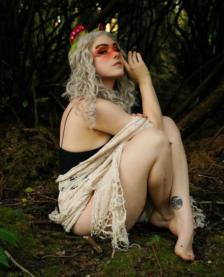 a woman with white curly hair and mushroom ears is sitting on the mossy forest floor. she is wearing a black top and has a beige shawl wrapped around her. she has an eye shadow make up on her eyes and a tattoo on her leg. the forest is visible in the background.