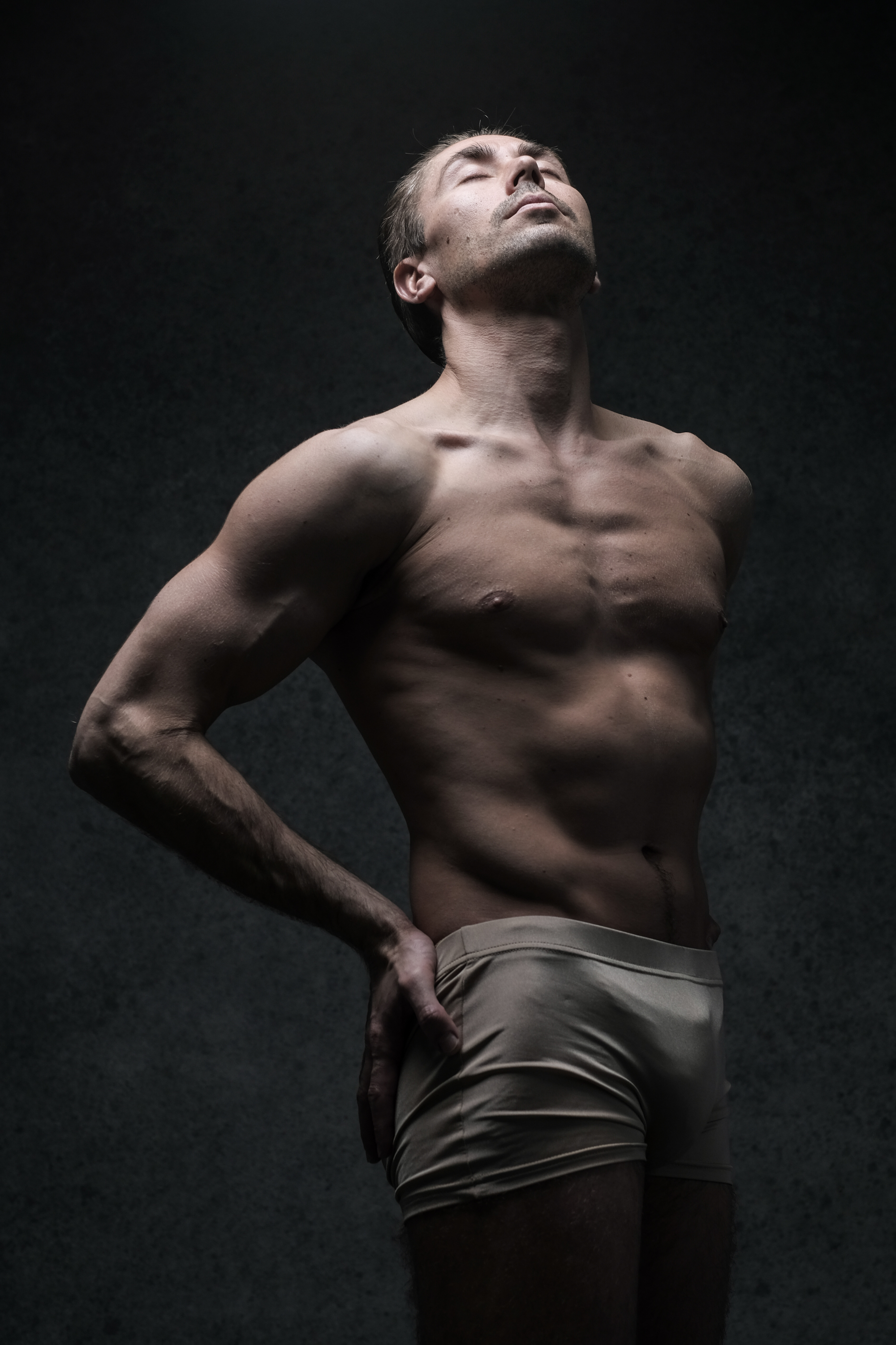 a muscular man stands in front of a dark gray background. he is wearing only underwear and is illuminated by a spotlight. his eyes are closed and his head is tilted back. his left hand is on his hip. the image is black and white.