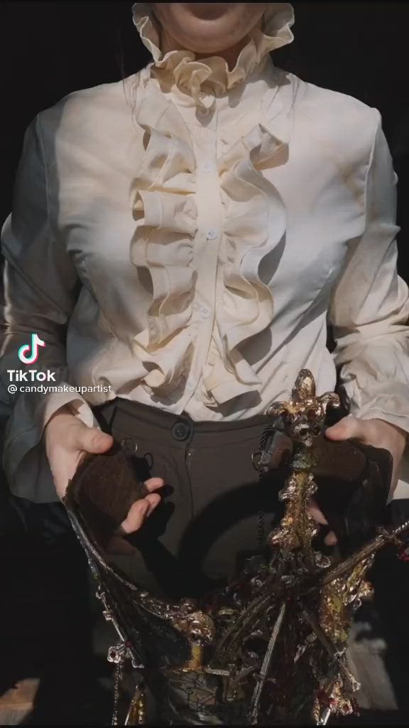 a person wearing a creamcolored ruffle shirt and dark pants holds a decorative, steampunkstyle object in their gloved hands.