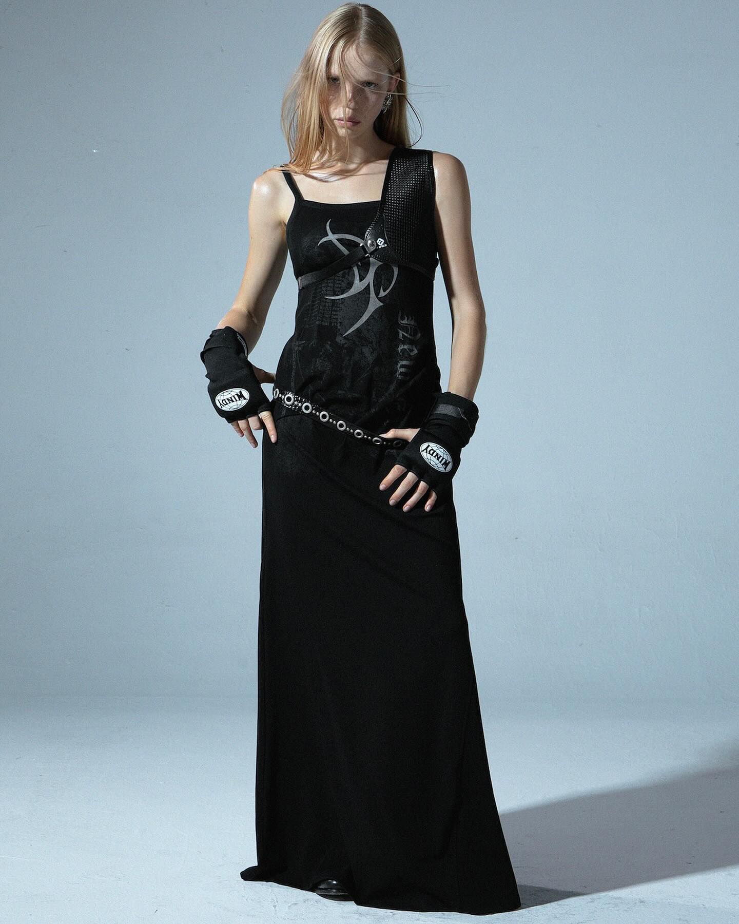 a fulllength shot of a young woman model in a black maxi dress with a tribalinspired design. she's wearing black hand wraps and a chain belt, and her pose is slightly edgy. the background is a simple, neutral grey. the overall style is goth or grunge inspired.
