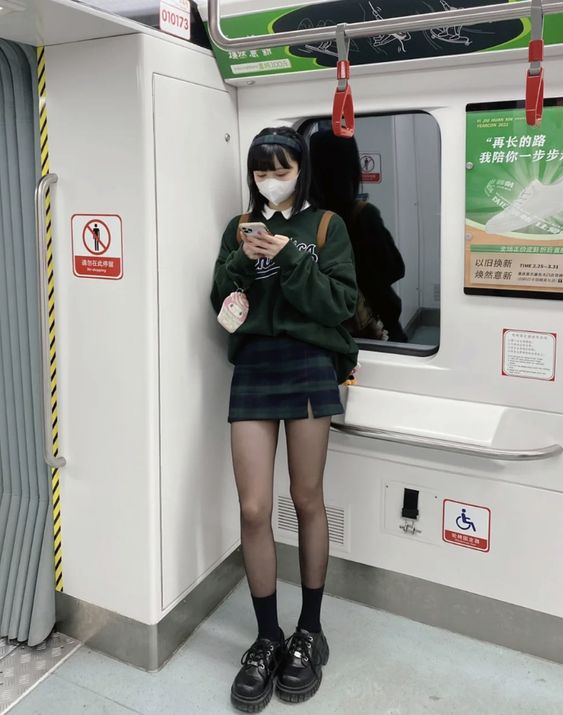 a young woman, wearing a green sweater, plaid skirt, black tights, and black shoes, stands in a subway car. she is looking down at her phone, and wearing a white face mask. the subway car is mostly empty, with only a few other people visible in the background. the lighting is bright and artificial. the overall impression is of a stylish and modern young woman going about her day.