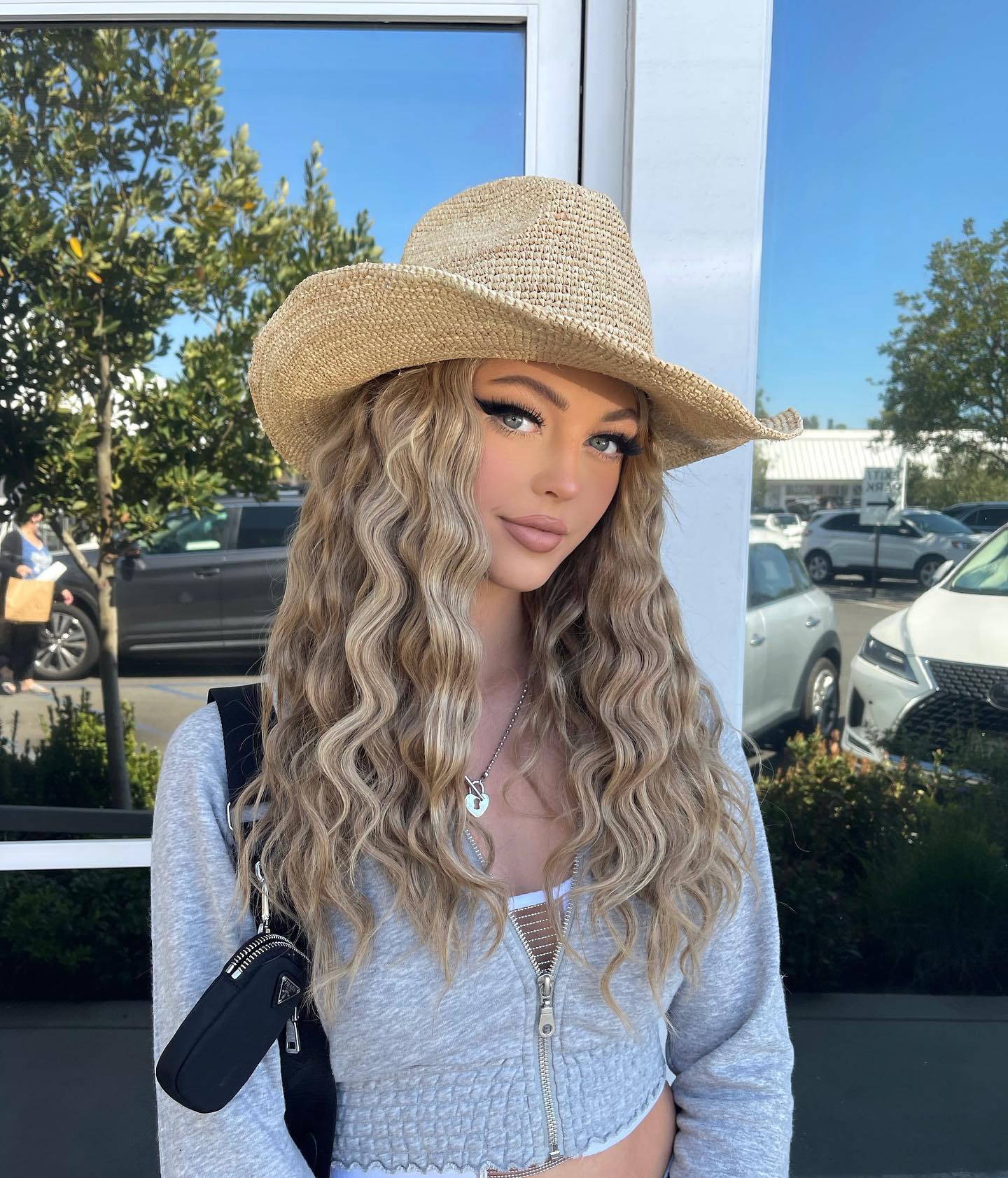 a woman is wearing a straw hat and a gray jacket with a purse, with long blonde curly hair and a silver necklace, standing outdoors in front of a building with cars and trees in the background.