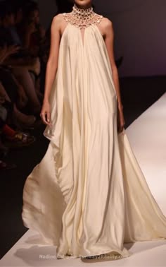 a beautiful woman in a white, long, flowing gown. she is on a runway and she is striking a pose for the camera. her hair is styled in a sleek and elegant way, and she is wearing a pearl necklace with a large center pendant. her makeup is flawless, and she looks incredibly confident. the gown is  draped and flowing, and it is the perfect example of how to dress for a special occasion. she is radiating a sense of glamour and sophistication.