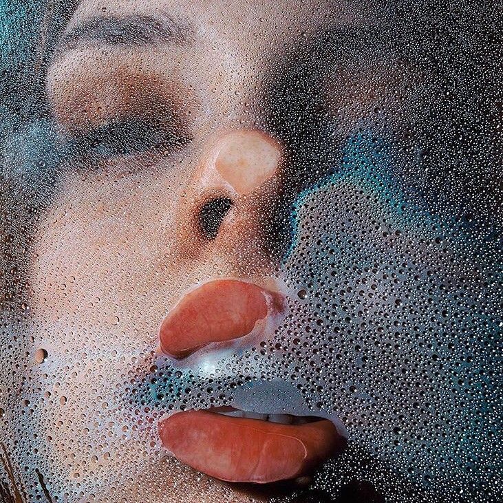 the image is a closeup, stylized portrait of a woman's face viewed through a surface covered in condensation or water droplets. the droplets create a blurry, diffused effect, partially obscuring the woman's features. her lips are full and painted a reddishbrown shade, and her eyes are partially visible with dark eye makeup. the overall mood is sensual, mysterious, and artistic, suggesting a theme of beauty or fantasy. the texture of the water droplets contrasts with the smoothness of the woman's skin. the color palette is muted and subdued, with a predominance of cool tones from the background contrasting with the warm tones of the woman’s face.