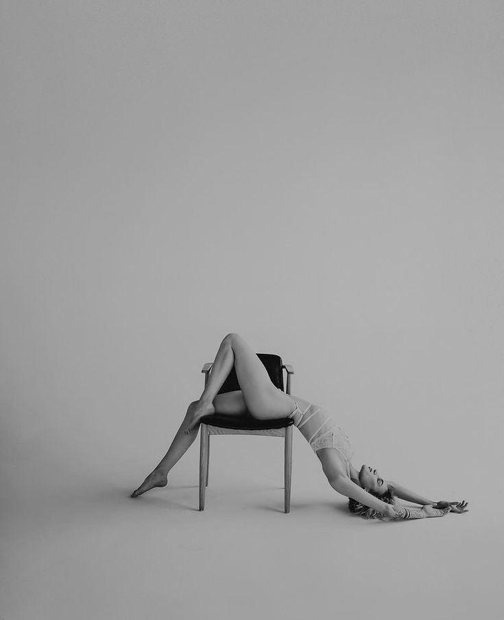 a black and white studio shot features a woman in a backbend pose, utilizing a chair. her legs are positioned on the chair, while her torso arches backwards, with her hands extended out on the floor. the image highlights her flexibility and contemporary style.
