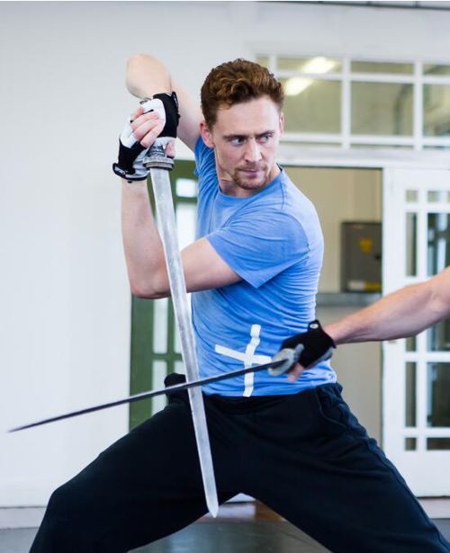 tom hiddleston, a renowned actor, is shown engaged in a rigorous fencing practice session. he stands poised, wearing black pants and a blue shirt, wielding a sword in his right hand. the intensity in his gaze and the sharpness of his movements reflect the seriousness of his training. the image captures the actor's dedication to the art of sword fighting, emphasizing his commitment to portraying a character authentically.