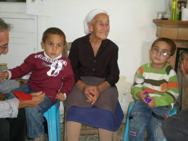 an elderly woman wearing a headscarf sits between two young boys.  the woman is smiling warmly. the boys are also sitting, and one is wearing a maroon sweatshirt while the other wears a striped longsleeved shirt. they appear to be in a simple, possibly rural home. the scene depicts a strong sense of family and generational connection.