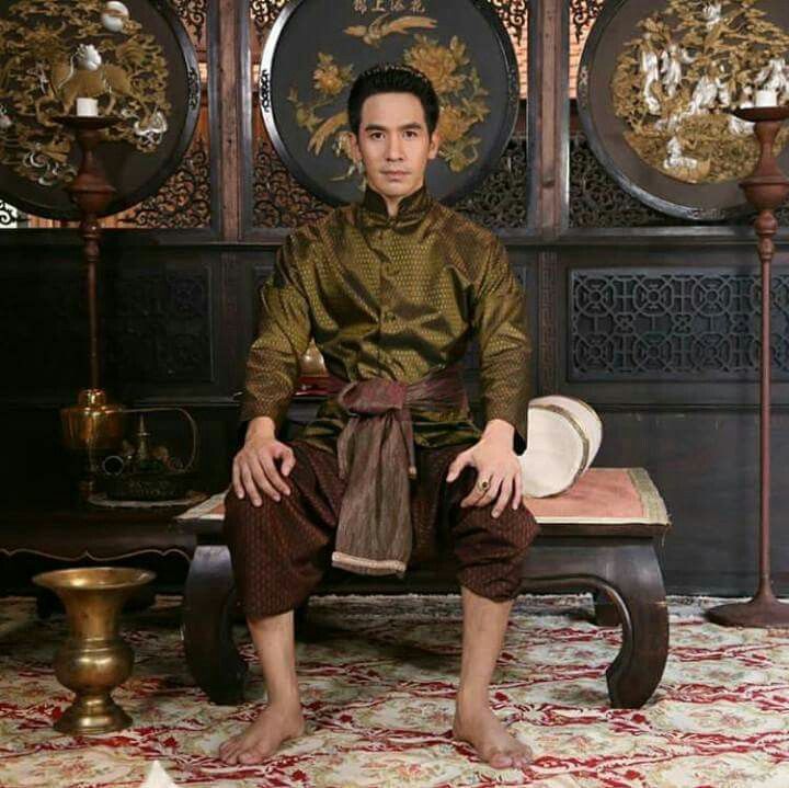 a man wearing traditional thai clothing is sitting on a vintage couch in a home. he is wearing a longsleeved,  goldtoned shirt with a sash around his waist. the man is wearing brown pants that match his sash. behind him, are two large, ornate, antique screens, with other decorative elements that match.