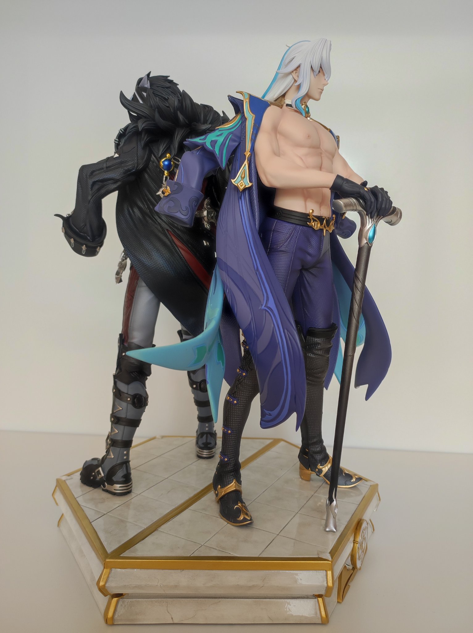 two anime figurines are standing on a white and gold platform base. the figurine on the left is dressed in black, with a black and blue cape and long boots. he has white hair, a sword, and a large, black hat. the figurine on the right is dressed in purple and white, with a long blue cape. he has white hair and is holding a long staff with a gold top. both figures are facing forward, and they are posed as though they are in a battle stance. the figurine on the left has a serious expression, while the figurine on the right has a determined expression. they are both standing on a white platform base with gold accents. this is a beautifully detailed figurine that would be a great addition to any anime fan's collection.