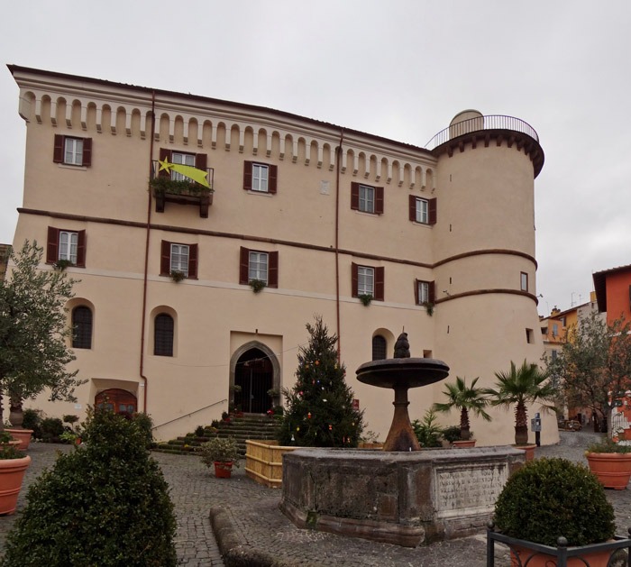 the image shows a large, light beige building with a medieval architecture style, possibly a castle or a historical building, featuring several windows with dark red shutters. a tower extends from one side of the building, with a rooftop railing. a bright yellow star is hung on a balcony. in the foreground, there's a stone fountain surrounded by plants, including an olive tree and a christmas tree decorated with lights. the ground is covered with cobblestones. the sky is overcast.