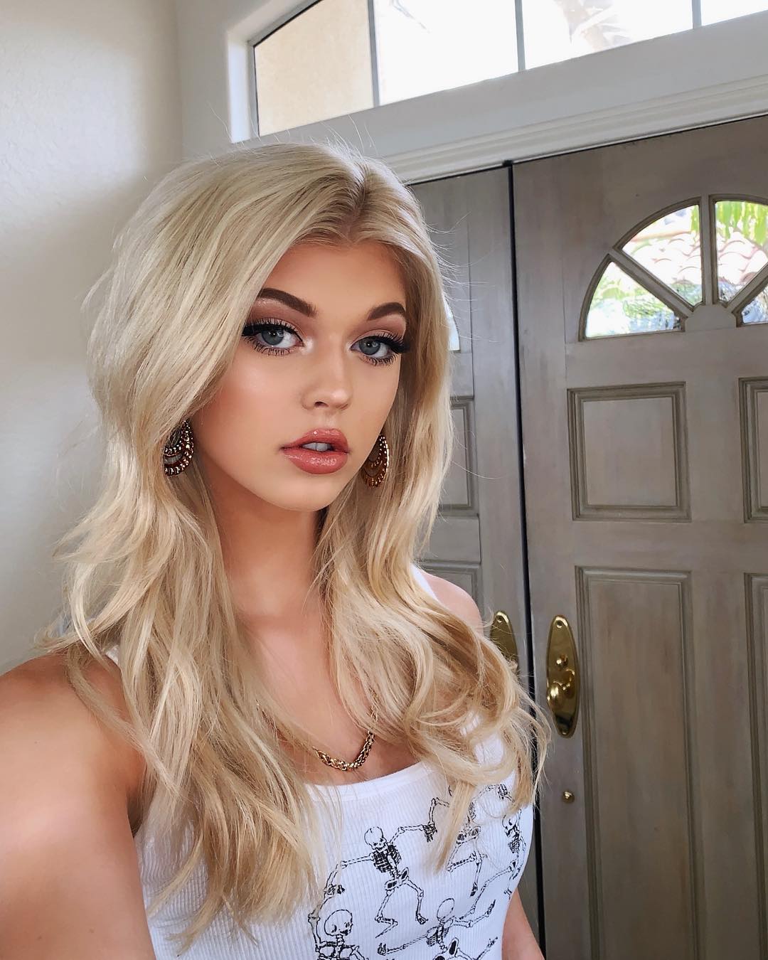 a portrait selfie of a woman with blonde hair and blue eyes wearing a white tank top with a skeleton print, earrings, and a necklace. she is standing in front of a closed door with a window above it.