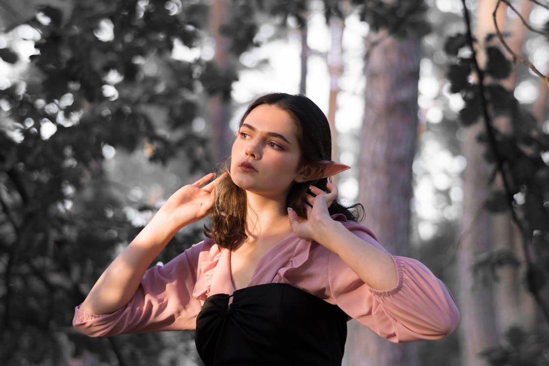 a young woman dressed as an elf stands in a forest, wearing a pink top with ruffles and a black bodice. she has pointed elf ears and is holding her hands near her face. the background is a forest with trees and blurred foliage, creating a fantasylike setting.