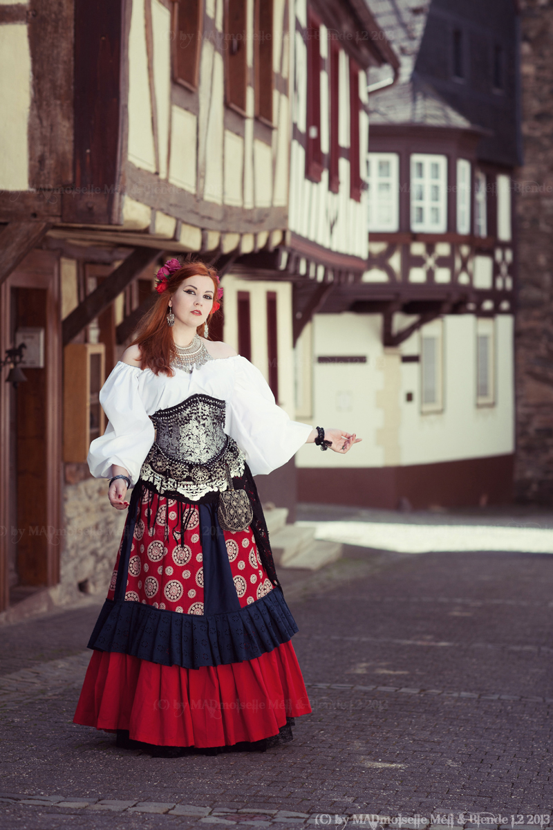 a woman in a traditional costume stands on a cobblestone street. she has long red hair with flowers in it, and is wearing a white blouse with long sleeves, a silver corset, and a red, blue and black skirt. the buildings behind her are halftimbered houses, with red and brown beams. there is also a stone building with a small window in the background. she is holding her arm out to the side and wearing a bracelet on her wrist. she also has a small purse hanging from her waist.