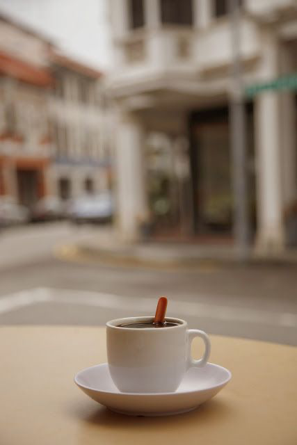 a white coffee cup with a brown coffee sits on a white saucer on a wooden table. the cup is sitting on a table with a blurry background of an urban street scene.