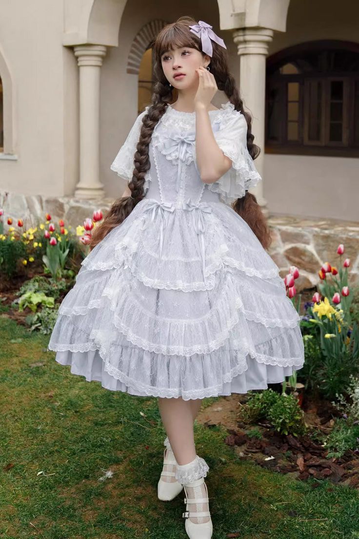 a young woman wearing a light blue and white lolita fashion dress is standing in a garden. she is posing for the camera with her hand on her face and a slight smile. the dress is made of lace and tulle, with layers of ruffles and a bow at the waist. she is also wearing white tights, white shoes, and a purple bow in her hair. the background features colorful tulips and daffodils, creating a soft and whimsical atmosphere. the natural lighting enhances the delicate colors and textures of the scene.