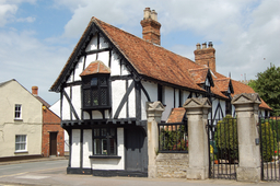 the image showcases a charming, historic tudorstyle house with its distinctive black and white halftimbered facade. the house features a steeply pitched roof covered in brown tiles and adorned with multiple chimneys. an oriel window, painted white, projects from the second floor, adding character to the building. black trim outlines the white walls and emphasizes the timber framing. a black iron gate, flanked by stone pillars, encloses the front yard, providing a glimpse of the greenery within. the house stands on a street corner, with another building partially visible in the background. a bright blue sky dotted with fluffy white clouds completes the scene.