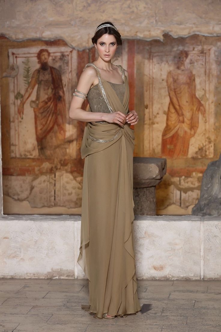 a fulllength shot of a female model wearing a flowing, beige grecianstyle gown. the dress is draped and features a cowl neckline. she is wearing a headband and subtle jewelry. the background is an ancient romanstyle setting with wall paintings depicting figures. the overall mood is elegant and sophisticated.