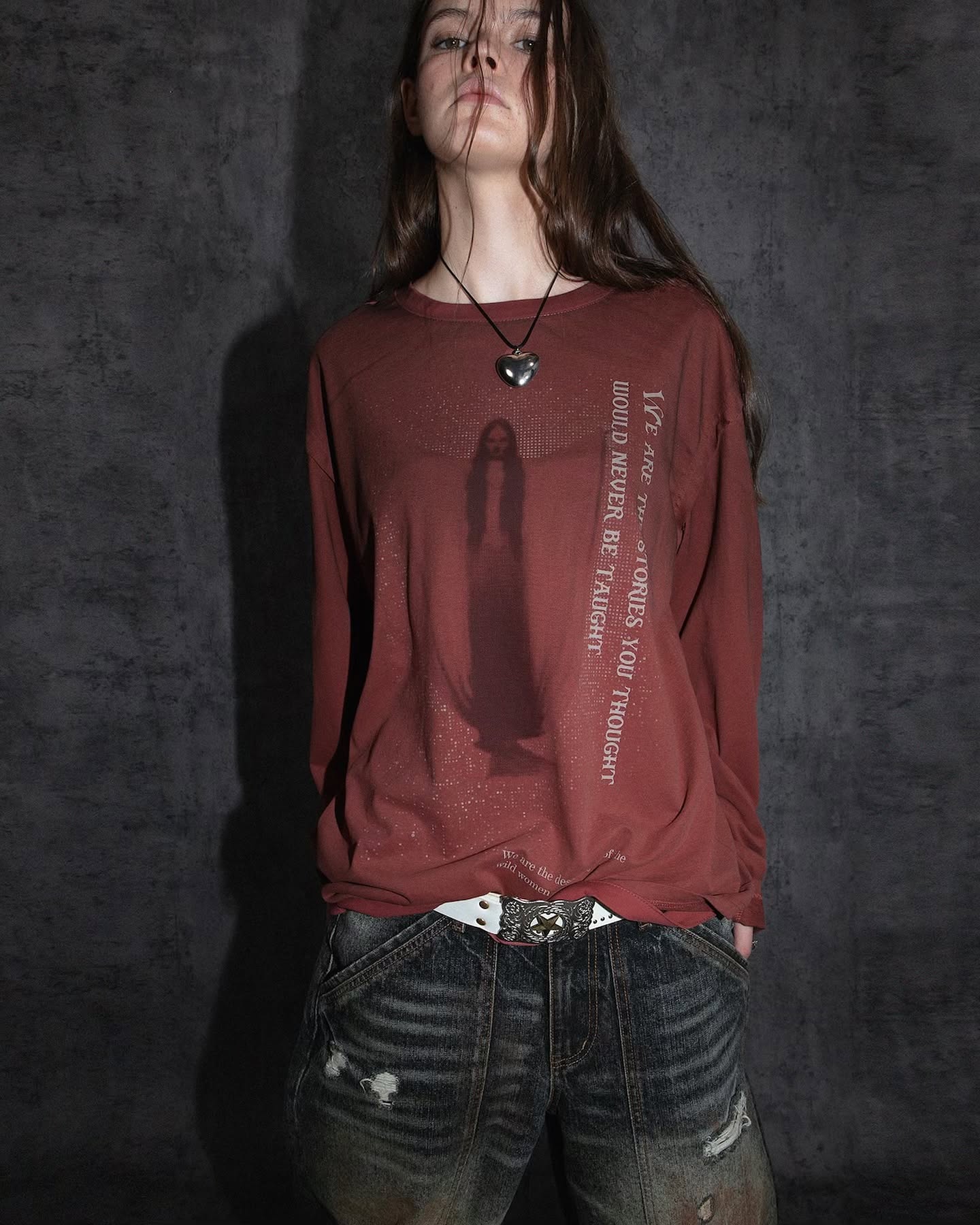 a full shot of a young woman wearing a longsleeve, maroon tshirt with a graphic print and text on the side. the tshirt is styled over distressed denim jeans. the woman wears a heartshaped necklace and a thin belt with metal accents. her dark brown hair is loose. the background is a dark muted grey. the overall mood of the image is dark, moody, and edgy, suggesting a fall/autumn fashion style. the model's pose is relaxed and casual.
