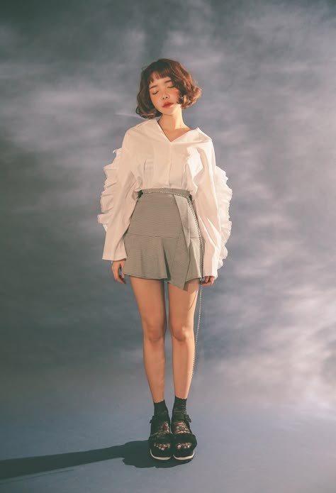 a young woman is standing in a studio with a cloudy background wearing a white ruffled blouse with a grey short skirt and black lace sandals. she has her eyes closed and is looking down. the image is welllit and the colors are muted.